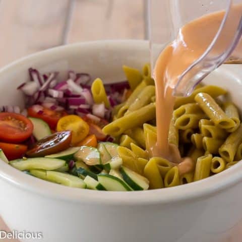 pouring homemade greek dressing from blender into large white bowl with gluten free green lentil penne, diced red onion, halved cherry tomatoes, and sliced cucumber