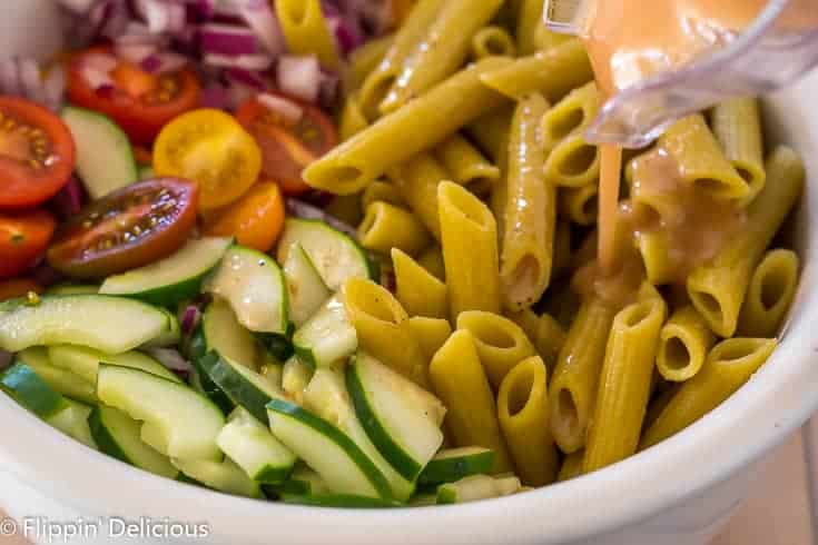 pouring homemade greek dressing from blender into large white bowl with gluten free green lentil penne, diced red onion, halved cherry tomatoes, and sliced cucumber