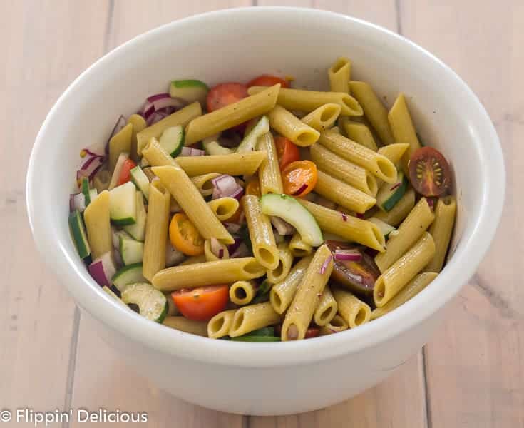 large white bowl with gluten free green lentil penne, diced red onion, halved cherry tomatoes, and sliced cucumber