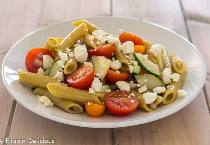 white plate with gluten free green lentil penne, diced red onion, halved cherry tomatoes, sliced cucumber, and feta cheese on a pale wood table