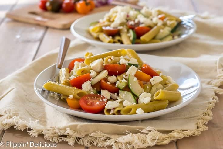 two white plates with gluten free greek pasta with cherry tomatoes, sliced cucumber, red onion, and feta on top of cream napkin on pale wood table with red onion and cherry tomatoes on cutting board in the background