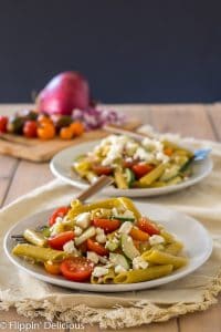 two white plates with gluten free greek pasta with cherry tomatoes, sliced cucumber, red onion, and feta on top of cream napkin on pale wood table with red onion and cherry tomatoes on cutting board in the background