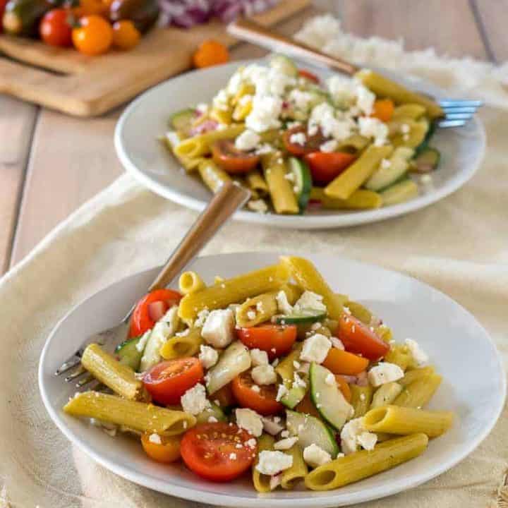 two white plates with gluten free greek pasta with cherry tomatoes, sliced cucumber, red onion, and feta on top of cream napkin on pale wood table with red onion and cherry tomatoes on cutting board in the background