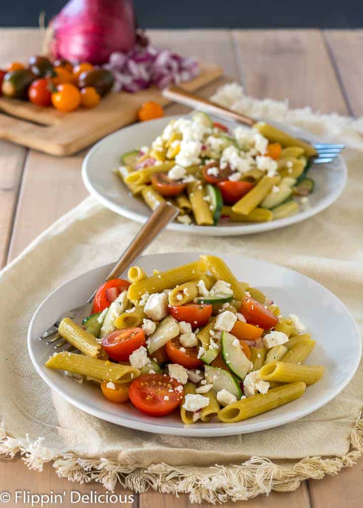 two white plates with gluten free greek pasta with cherry tomatoes, sliced cucumber, red onion, and feta on top of cream napkin on pale wood table with red onion and cherry tomatoes on cutting board in the background