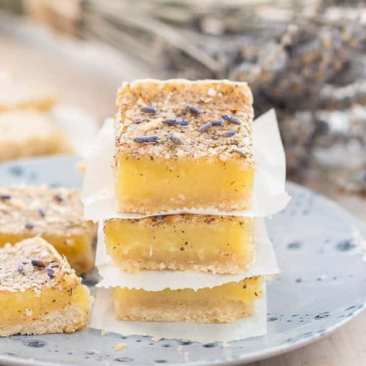 three gluten free lavender lemon bars stocked on top of each other, on a blue plate with two more lavender lemon bars beside them. Sprinkled with lavender buds and a bouquet of dried lavender in the background