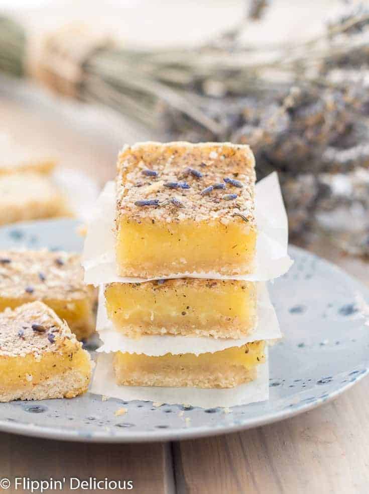 three gluten free lavender lemon bars stacked on top of each other, on a blue plate with two more lavender lemon bars beside them. Sprinkled with lavender buds and a bouquet of dried lavender in the background