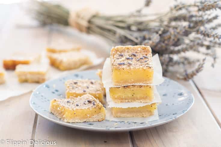 three gluten free lavender lemon bars stacked on top of each other, on a blue plate with two more lavender lemon bars beside them. Sprinkled with lavender buds and a bouquet of dried lavender in the background