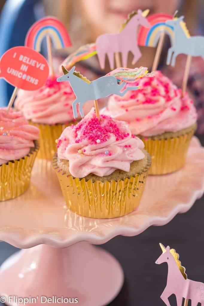 gluten free yellow cupcake in gold wrapper with pink vegan strawberry frosting and blue unicorn cupcake topper on a pink cake pedestal with more cupcakes in the background