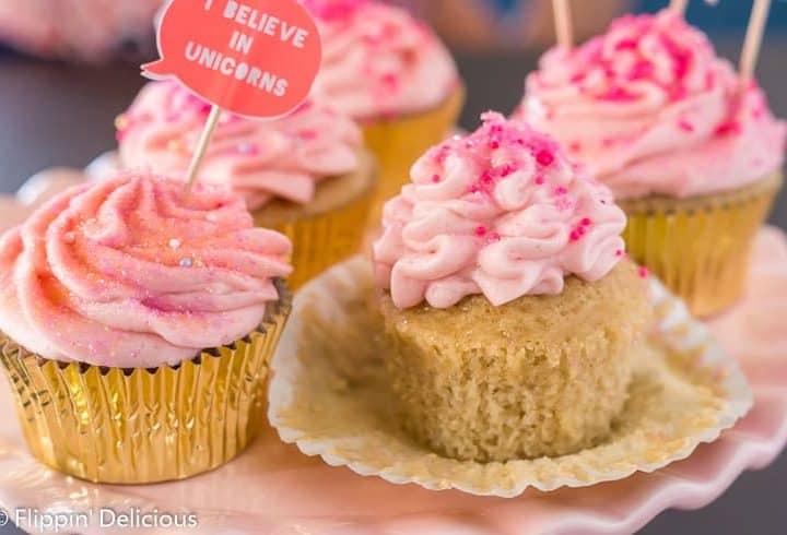 unwrapped gluten free yellow cupcake topped with pink strawberry frosting and pink sprinkles, on a pink cake pedestal with more pink frosted cupcakes in gold wrappers in the background