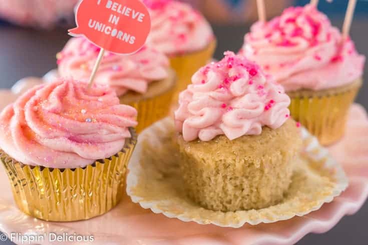 unwrapped gluten free yellow cupcake topped with pink strawberry frosting and pink sprinkles, on a pink cake pedestal with more pink frosted cupcakes in gold wrappers in the background