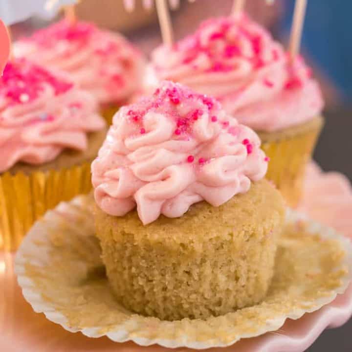 unwrapped gluten free yellow cupcake topped with pink strawberry frosting and pink sprinkles, on a pink cake pedestal with more pink frosted cupcakes in gold wrappers and unicorn and rainbow cake toppers in the background