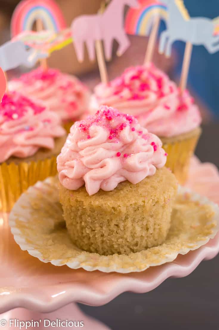 unwrapped gluten free yellow cupcake topped with pink strawberry frosting and pink sprinkles, on a pink cake pedestal with more pink frosted cupcakes in gold wrappers and unicorn and rainbow cake toppers in the background