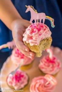 girl in blue dress holding a gluten free yellow cupcake with pink strawberry frosting, pink non pareil sprinkles, and a pink unicorn paper cupcake topper over a pink cake plate with four more pink frosted cupcakes