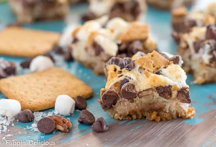 vegan gluten free magic cookie bars with marshallows, pecans, coconut, graham crackers, and chocolate chips on a teal and gray table