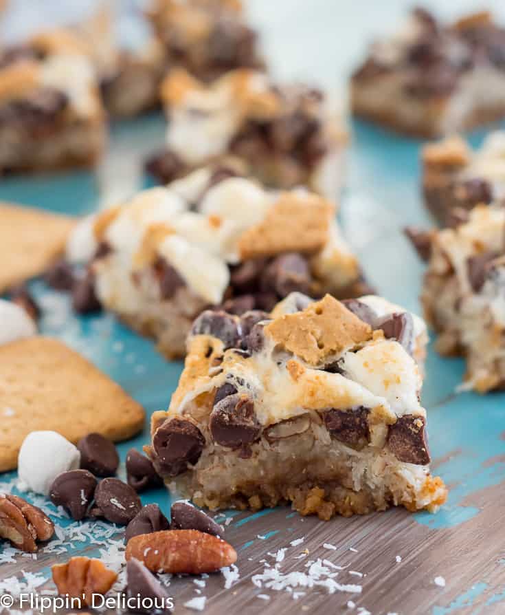 vegan gluten free magic cookie bars with marshmallows on a teal and gray table with schar honeygrams, pecans, chocolate chips, and marshallows 