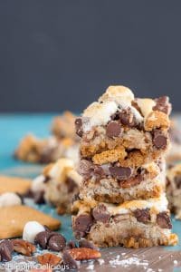 stack of three vegan gluten free magic cookie bars with marshallows, pecans, coconut, graham crackers, and chocolate chips on a teal and gray table