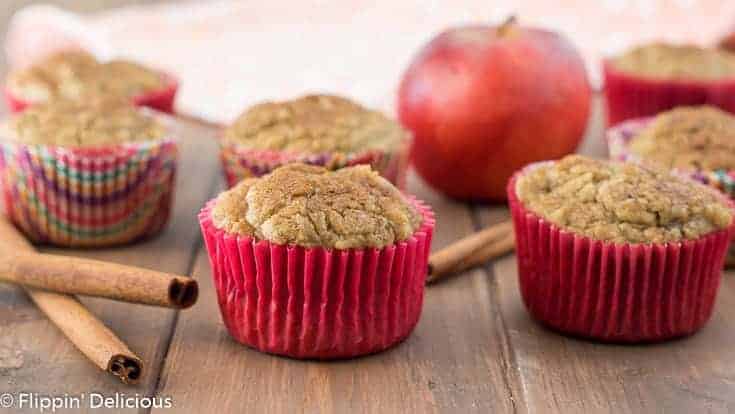 gluten free apple cinnamon muffins on a wooden table with cinnamon sticks, an apple and a dish towel in the background 