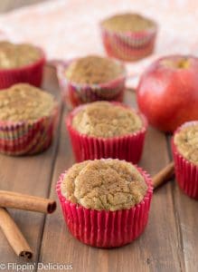 gluten free apple cinnamon muffins on a wooden table with cinnamon sticks, an apple and a dish towel in the background