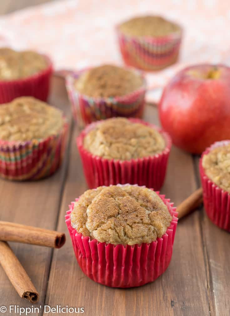 gluten free apple cinnamon muffins on a wooden table with cinnamon sticks, an apple and a dish towel in the background 