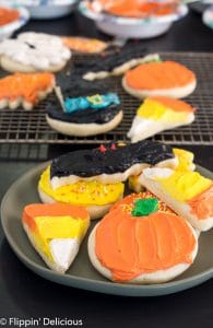 frosted gluten free Halloween sugar cookies including two candy corn sugar cookies, a pumpkin sugar cookie, a bat sugar cookie, and a witch hat sugar cookie, on a plate with more gluten free cut out cookies in the background