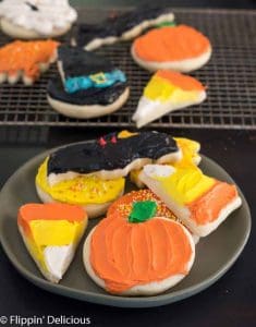 frosted gluten free Halloween sugar cookies including two candy corn sugar cookies, a pumpkin sugar cookie, a bat sugar cookie, fall leaf, and a witch hat sugar cookie, on a plate with more gluten free cut out cookies in the background