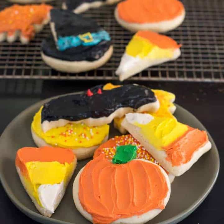 frosted gluten free Halloween sugar cookies including two candy corn sugar cookies, a pumpkin sugar cookie, a bat sugar cookie, fall leaf, and a witch hat sugar cookie, on a plate with more gluten free cut out cookies in the background