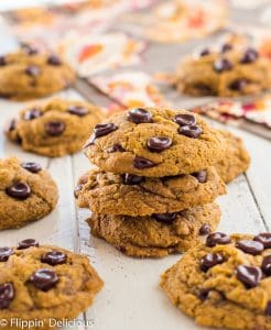 a stack of three gluten free vegan pumpkin chocolate chip cookies on a white wood table, surrounded by more gluten free pumpkin chocolate chip cookies with a orange and brown patterned napkin in the background