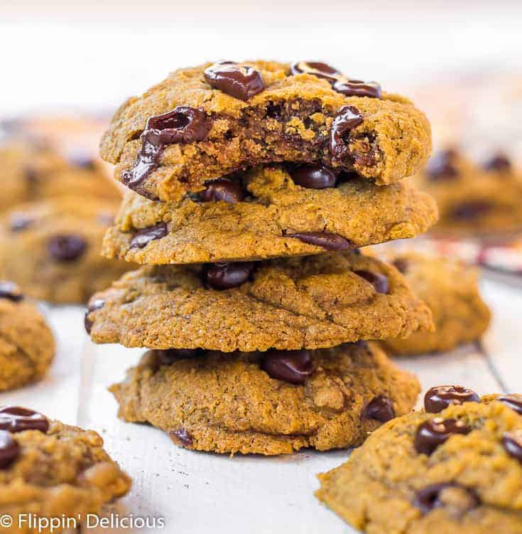 a stack of three gluten free vegan pumpkin chocolate chip cookies, with a bite taken out of the top pumpkin chocolate chip cookie on a white wood table, surrounded by more gluten free pumpkin chocolate chip cookies with a orange and brown patterned napkin in the background