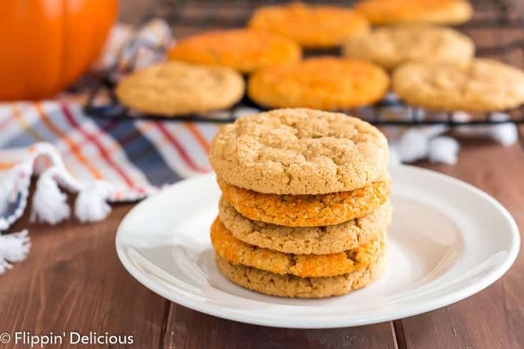 stack of five gluten free pumpkin sugar cookies on a white plate with a cooling rack with more vegan gluten free pumpkin sugar cookies, an orange and navy plaid kitchen towel, and pumpkin in the background