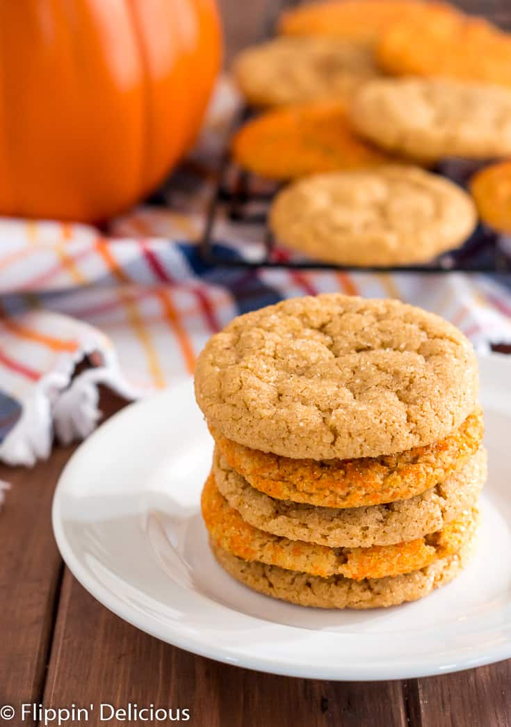 stack of five gluten free pumpkin sugar cookies on a white plate with a cooling rack with more vegan gluten free pumpkin sugar cookies, an orange and navy plaid kitchen towel, and pumpkin in the background