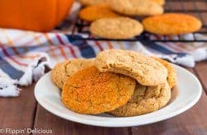 white plate with five vegan gluten free pumpkin sugar cookies with a bite taken out of the top cookie with a cooling rack with more vegan gluten free pumpkin sugar cookies, an orange and navy plaid kitchen towel, and pumpkin in the background