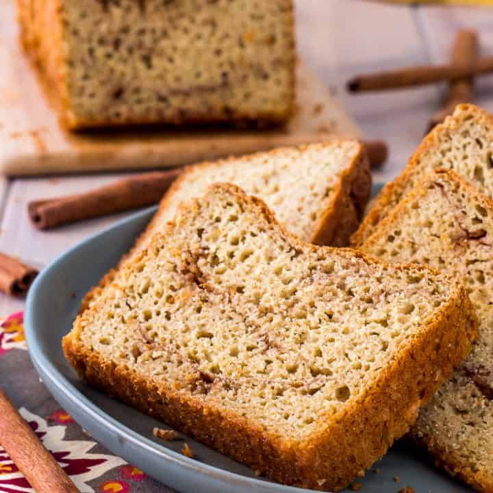 slices of gluten free cinnamon swirl bread on green plate placed on an orange patterned napkin with full loaf of gluten free cinnamon bread in the background.