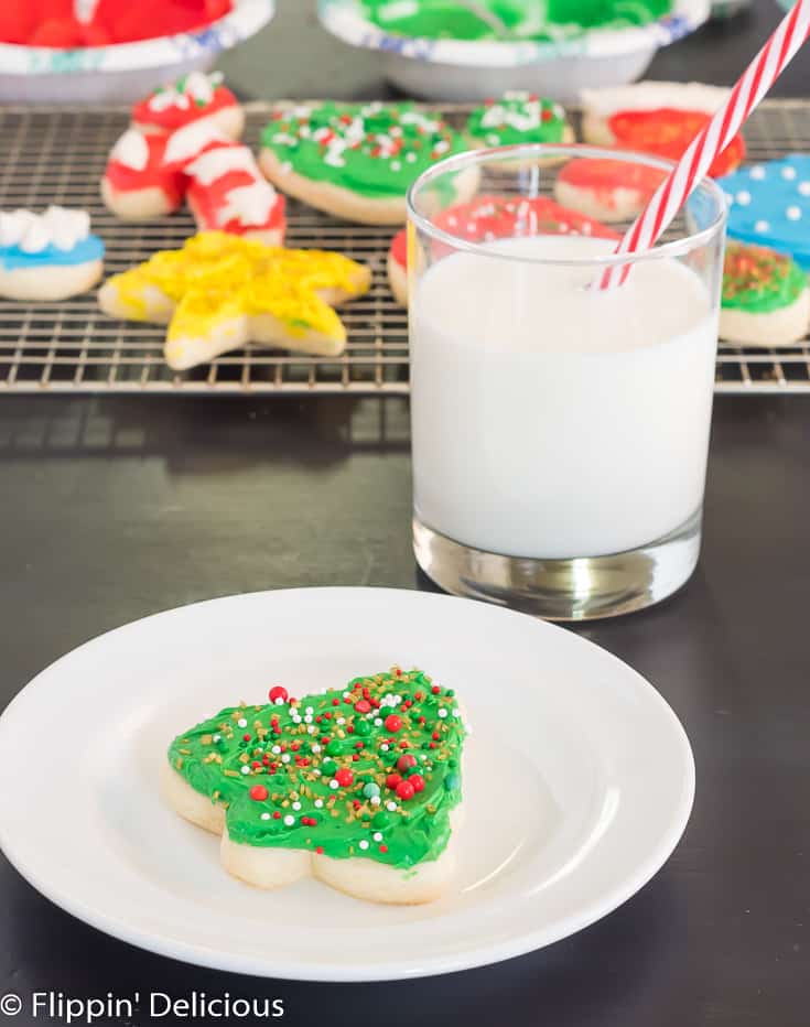 gluten free sugar cookie cutout shaped like a holiday tree with green frosting and sprinkles, on a white plate beside a glass of milk with a red and white striped straw with a cooling rack full of gluten free sugar cookie cutouts in the background
