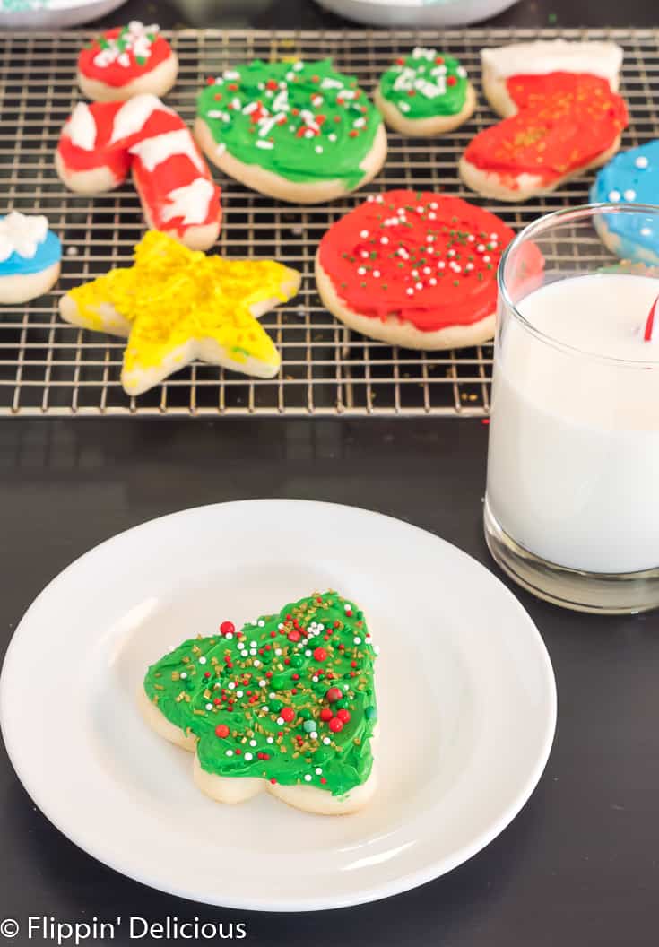 gluten free sugar cookie cutout shaped like a holiday tree with green frosting and sprinkles, on a white plate beside a glass of milk with a red and white striped straw with a cooling rack full of gluten free sugar cookie cutouts in the background