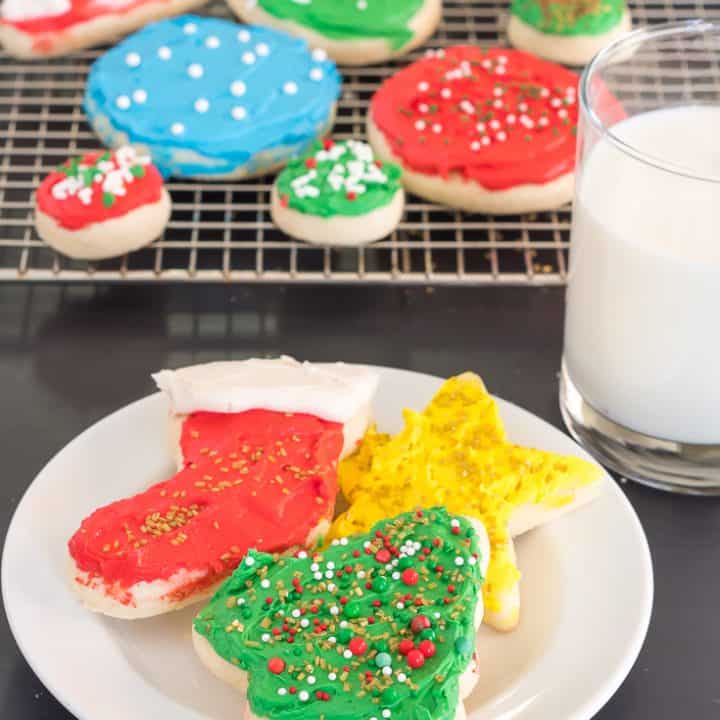 gluten free sugar cookie cutout shaped like a holiday tree, christmas stocking, and star, each topped with frosting and sprinkles, on a white plate beside a glass of milk with a red and white striped straw with a cooling rack full of gluten free sugar cookie cutouts in the background