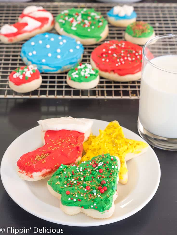gluten free sugar cookie cutout shaped like a holiday tree, christmas stocking, and star, each topped with frosting and sprinkles, on a white plate beside a glass of milk with a red and white striped straw with a cooling rack full of gluten free sugar cookie cutouts in the background