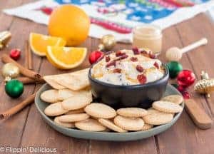 cranberry cream cheese dip drizzled with honey sprinkled with dried cranberries in black bowl on green plate filled with gluten free entertainment crackers on wooden table with sliced oranges, honey, and ornaments in the background