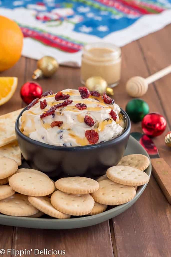 cranberry cream cheese dip drizzled with honey sprinkled with dried cranberries in black bowl on green plate filled with gluten free entertainment crackers on wooden table with sliced oranges, honey, and ornaments in the background