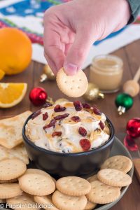 hand dipping gluten free schar entertainment cracker into cranberry cream cheese dip drizzled with honey sprinkled with dried cranberries in black bowl on green plate filled with gluten free entertainment crackers on wooden table with sliced oranges, honey, and ornaments in the background