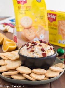 cranberry cream cheese dip drizzled with honey sprinkled with dried cranberries in black bowl on green plate filled with gluten free entertainment crackers on wooden table with sliced oranges, honey, and ornaments in the background