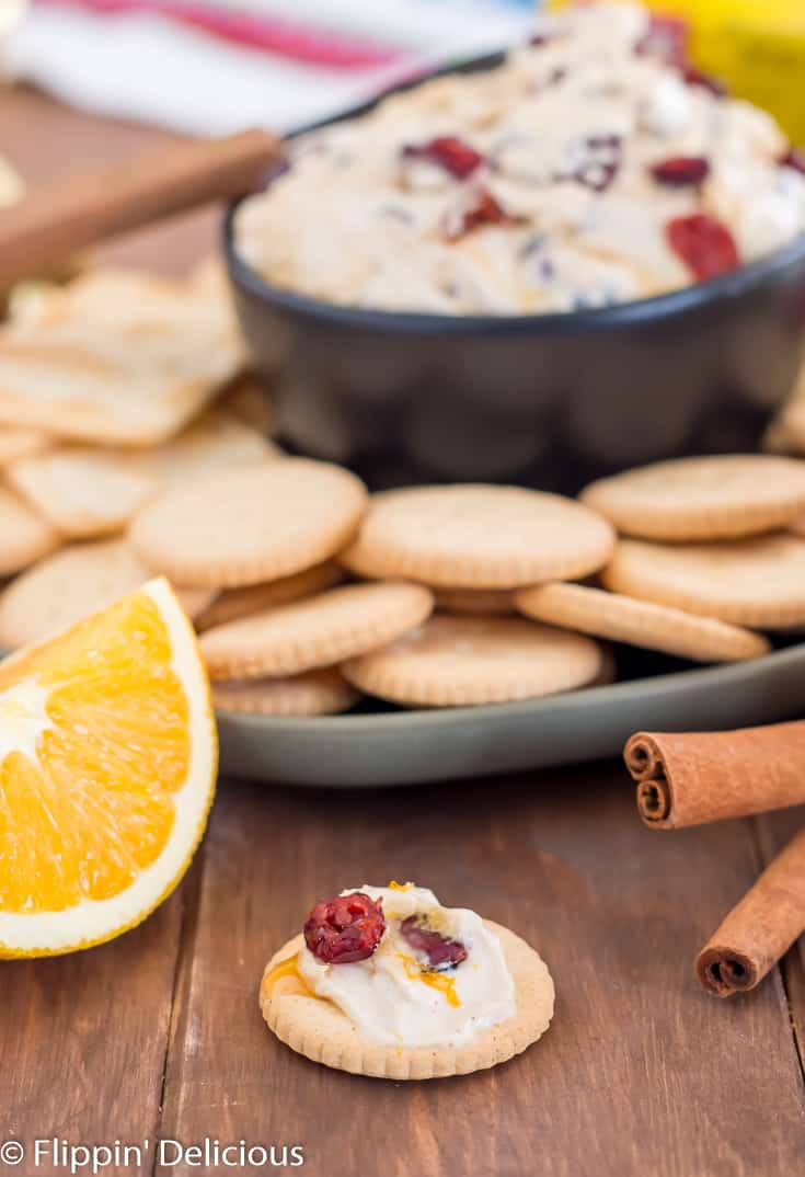 gluten free schar entertainment cracker topped with cranberry cream cheese dip drizzled with honey sprinkled with dried cranberries in black bowl on green plate filled with gluten free entertainment crackers on wooden table with sliced oranges, honey, and ornaments in the background