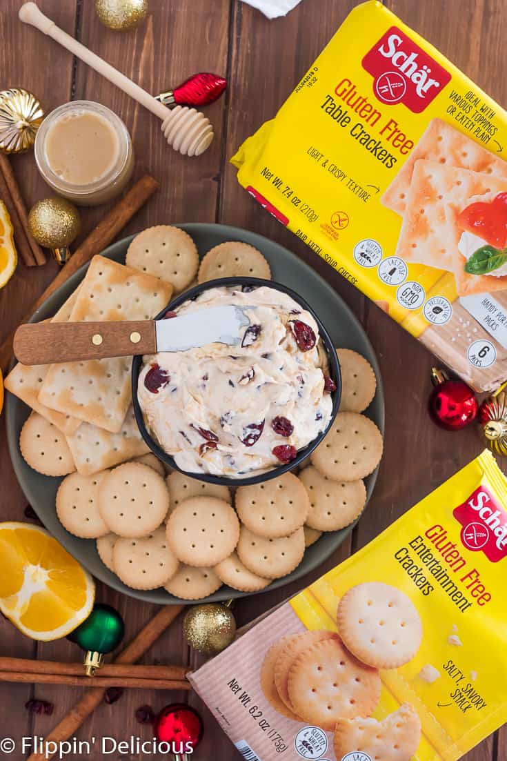 Overhead view of cranberry cream cheese dip drizzled with honey sprinkled with dried cranberries and spreader in black bowl on green plate filled with gluten free entertainment crackers on wooden table beside Schar Entertainment Crackers and Schar Table crackers packaging, sliced oranges, honey, and ornaments in the background