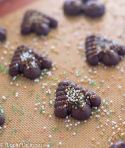 unbaked gluten free chocolate spritz cookies with white, gold, and green sprinkles on a silicone baking mat