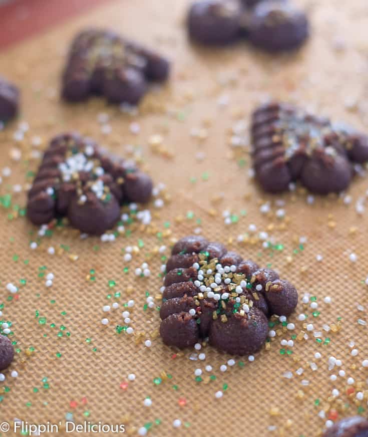 unbaked gluten free chocolate spritz cookies with white, gold, and green sprinkles on a silicone baking mat