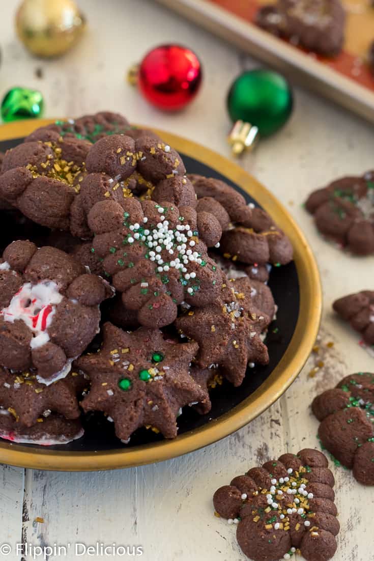 gluten free chocolate spritz cookies on a black plate rimmed with gold on a white farm house table surrounded by more gluten free chocolate spritz cookies and red, green, and gold christmas ornaments