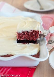 removing slice of dairy free gluten free red velvet sheet cake topped with white frosting from a white scallop-edged pan of cake in he background