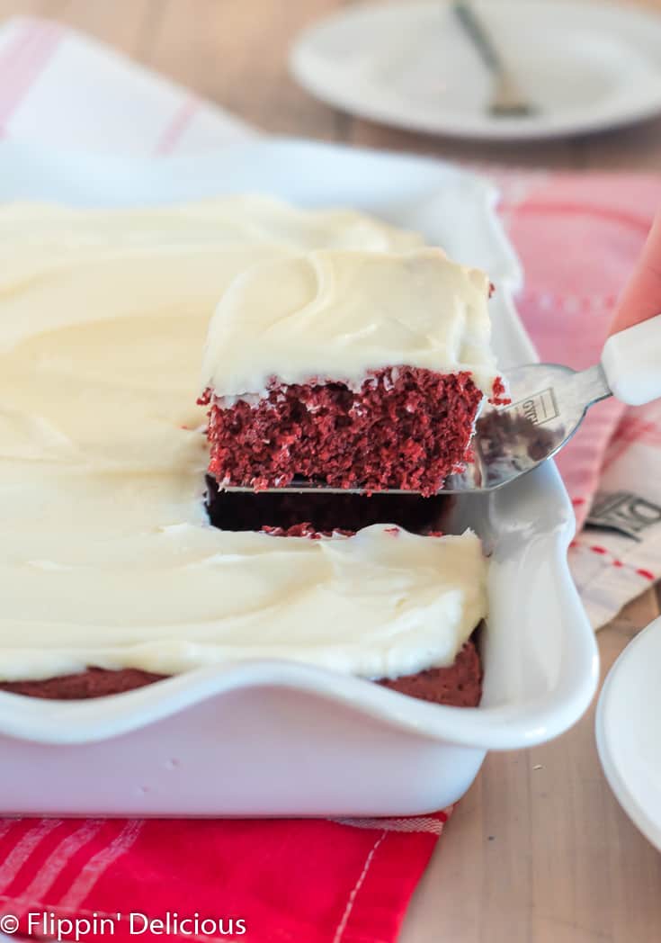 removing slice of dairy free red velvet sheet cake topped with white frosting from a white scallop-edged pan of cake in he background