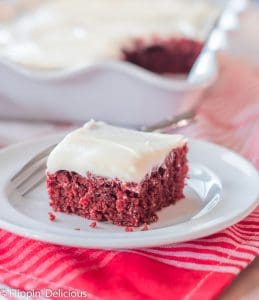 slice of dairy free gluten free red velvet sheet cake topped with white frosting on a white plate with a bite on a small fork with white scallop-edged pan of cake in the background