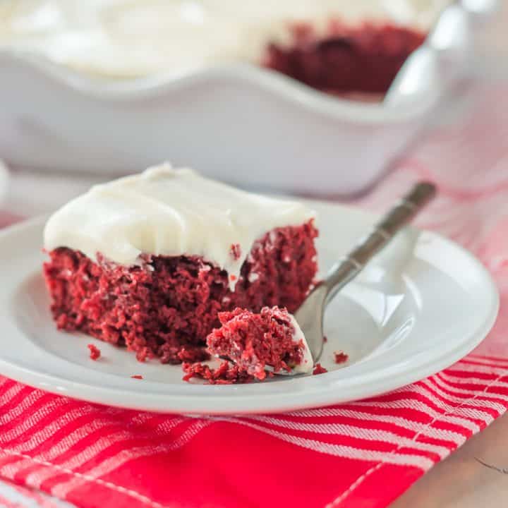 slice of dairy free gluten free red velvet sheet cake topped with white frosting on a white plate with a bite on a small fork with white scallop-edged pan of cake int he background