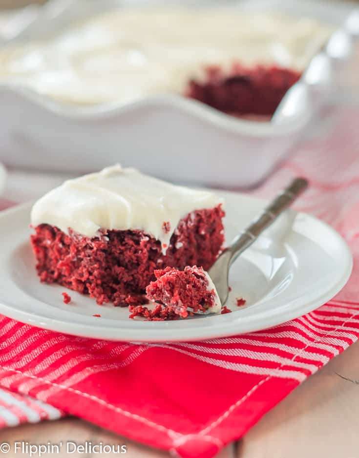 slice of dairy free red velvet sheet cake topped with white frosting on a white plate with a bite on a small fork with white scallop-edged pan of cake in the background